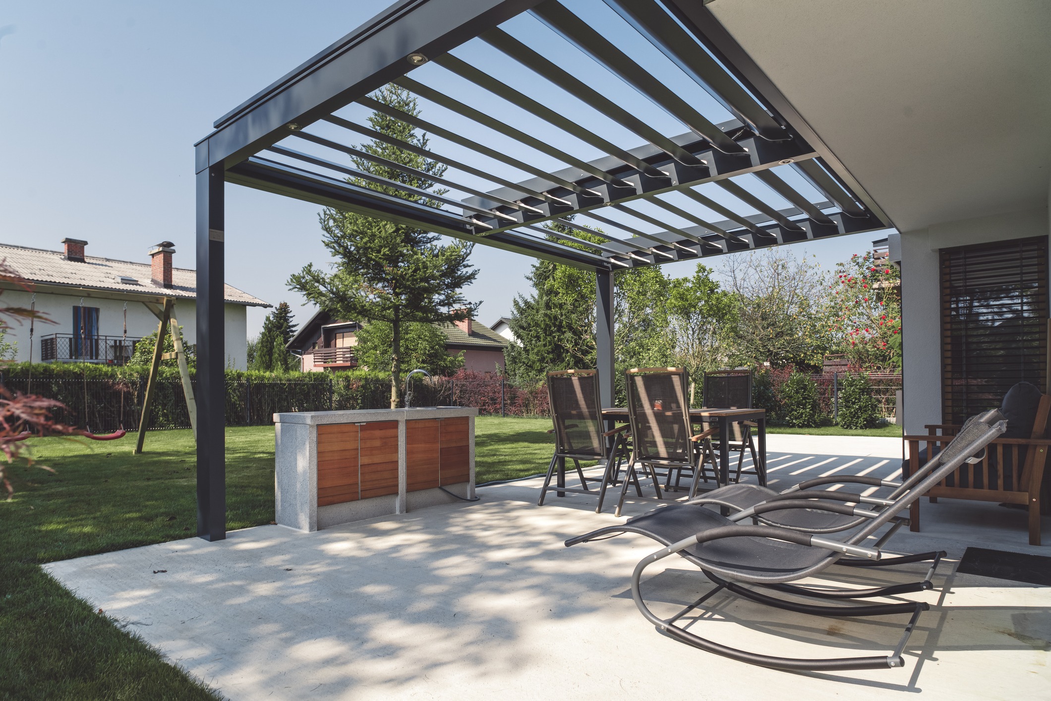 A modern backyard patio with a metal pergola, a bar counter with four high chairs, two black lounge chairs, and a wooden armchair. The patio overlooks a green lawn with trees and neighboring houses.