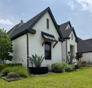 A modern white brick house with black trim features large windows, spiky agave plants in raised beds, and a well-maintained lawn under a partly cloudy sky. Shrubs and small trees border the house.