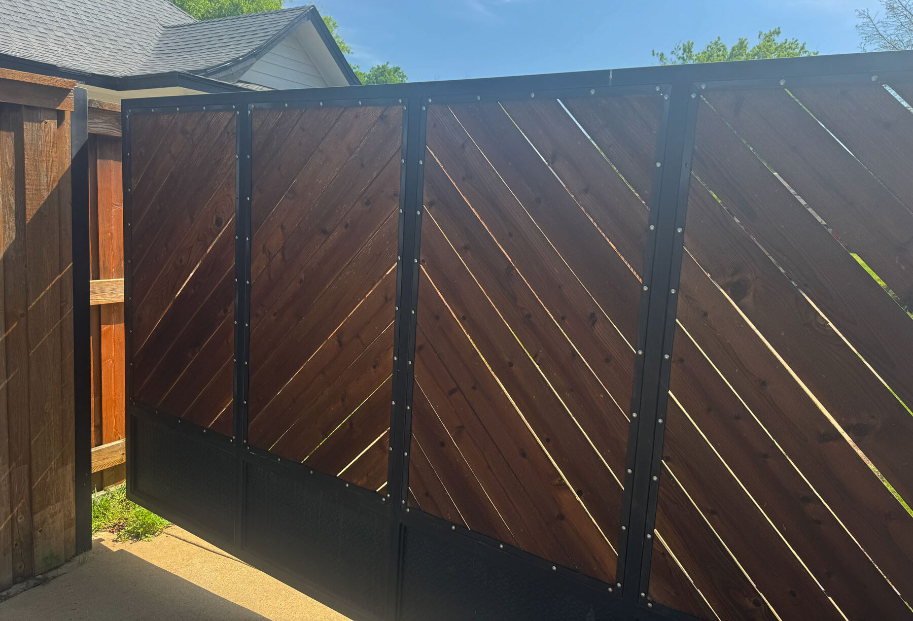 A modern wooden fence with diagonal planks and black metal framing stands between a house with a gray roof and another wooden fence, under a partly cloudy sky.
